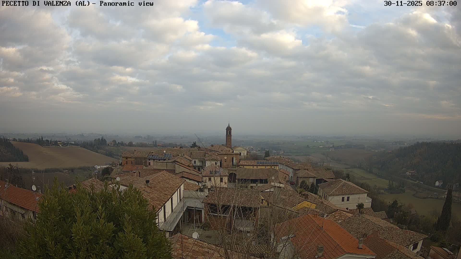 A panoramic view captures a hillside village with a prominent bell tower and terracotta rooftops, surrounded by rolling agricultural lands and distant hazy valleys, all beneath a partly cloudy and dull sky.