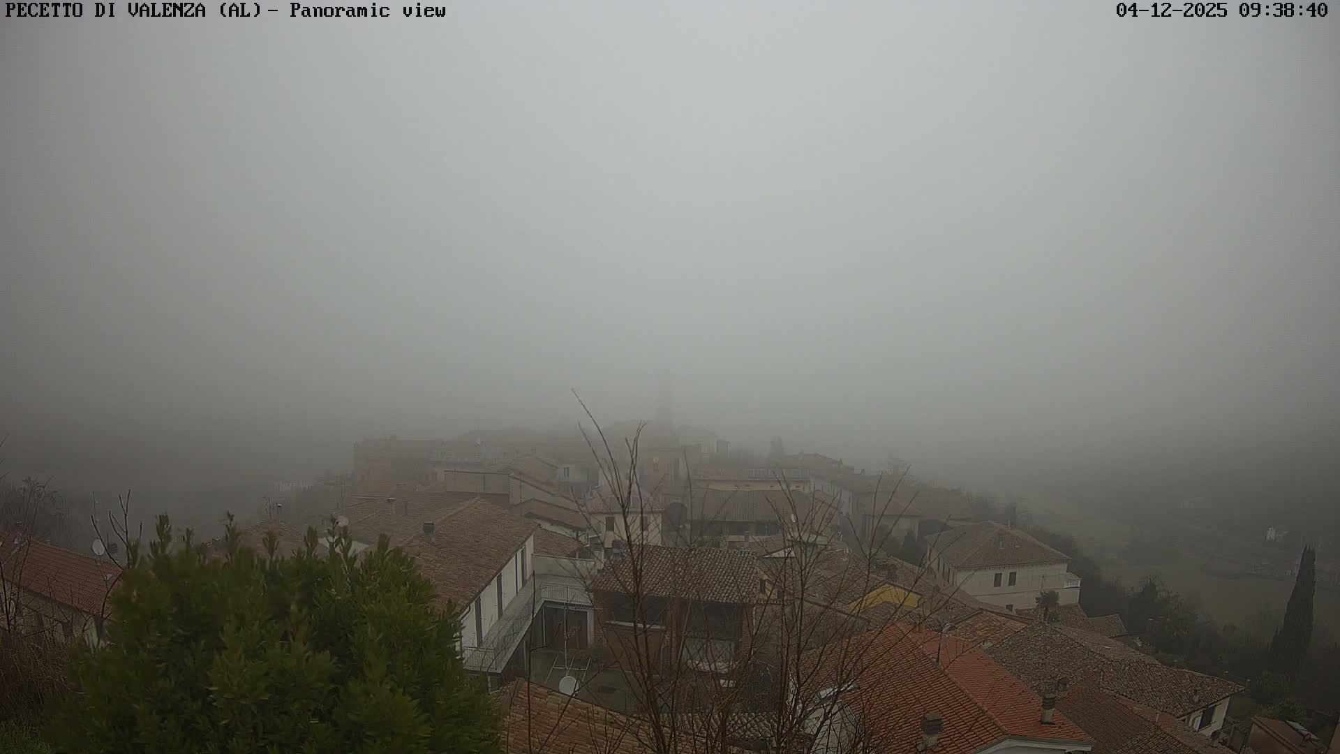 A panoramic view of a town with numerous buildings and red-tiled roofs is enveloped in thick fog, obscuring the distant landscape.