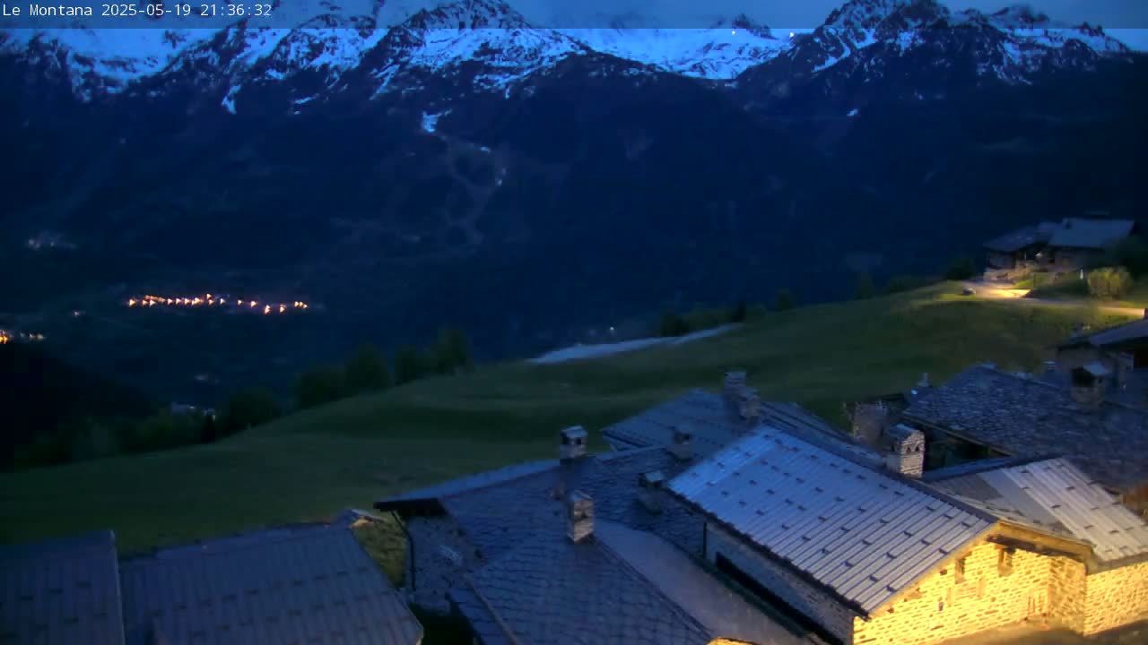 A nighttime view of a village nestled in a valley, with snow-capped mountains in the background under a dark, clear sky.
