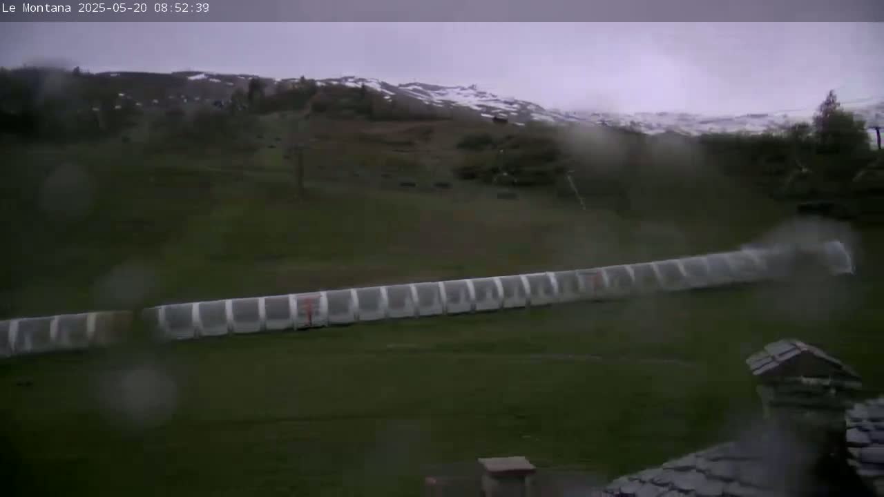 A long, covered structure sits on a grassy field, overlooking a snow-dusted hillside under a rainy, overcast sky.