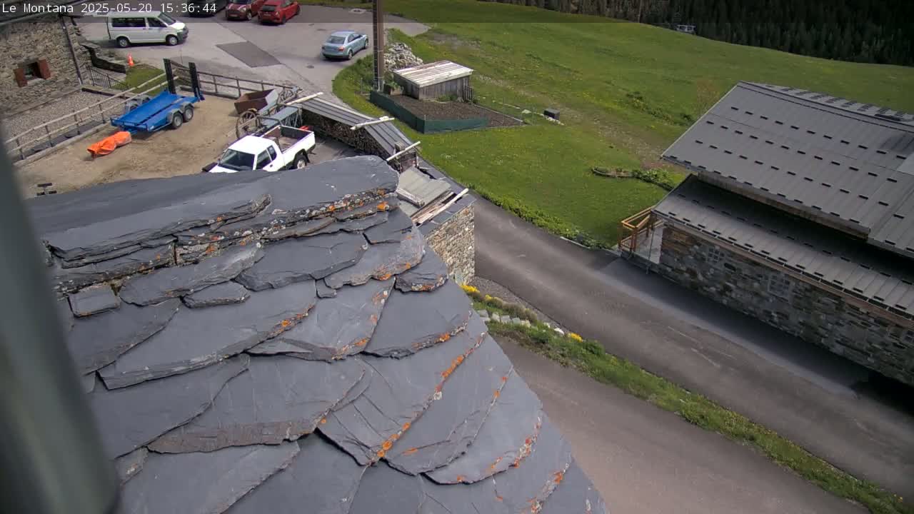 A high-angle view shows a slate roof with orange lichen, a parking area with several vehicles, and a grassy field under a sunny sky.