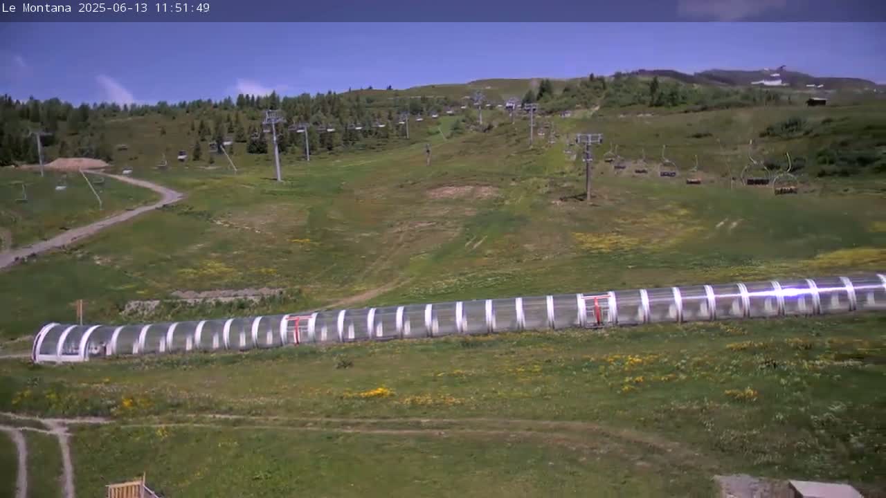 A long, clear-covered structure sits on a grassy hillside dotted with wildflowers under a mostly sunny sky, with several ski lifts visible in the background.