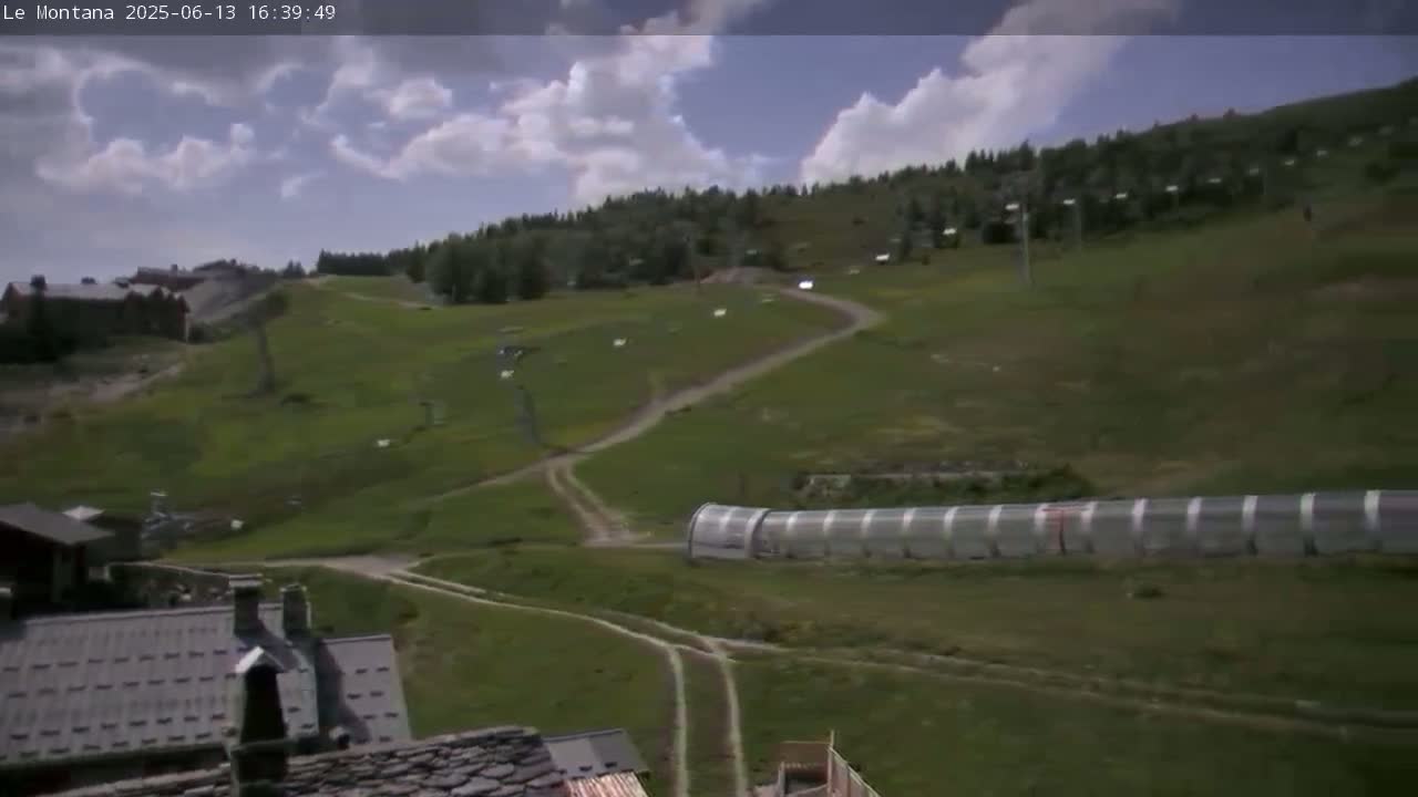 A partly cloudy day reveals a mountainous landscape with grassy hills, dirt roads, ski lift infrastructure, and buildings, including a long, covered structure.