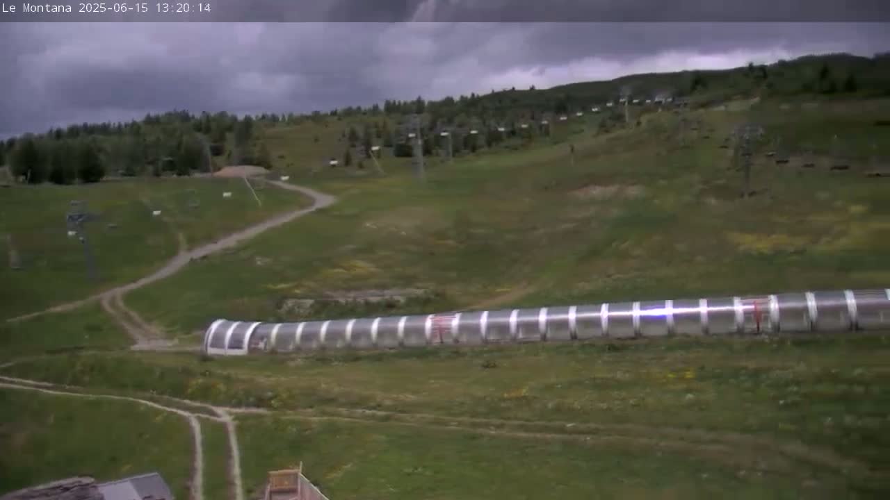 A long, translucent tunnel-like structure sits in a grassy field under an overcast sky, with several ski lift structures and dirt paths visible in the background.