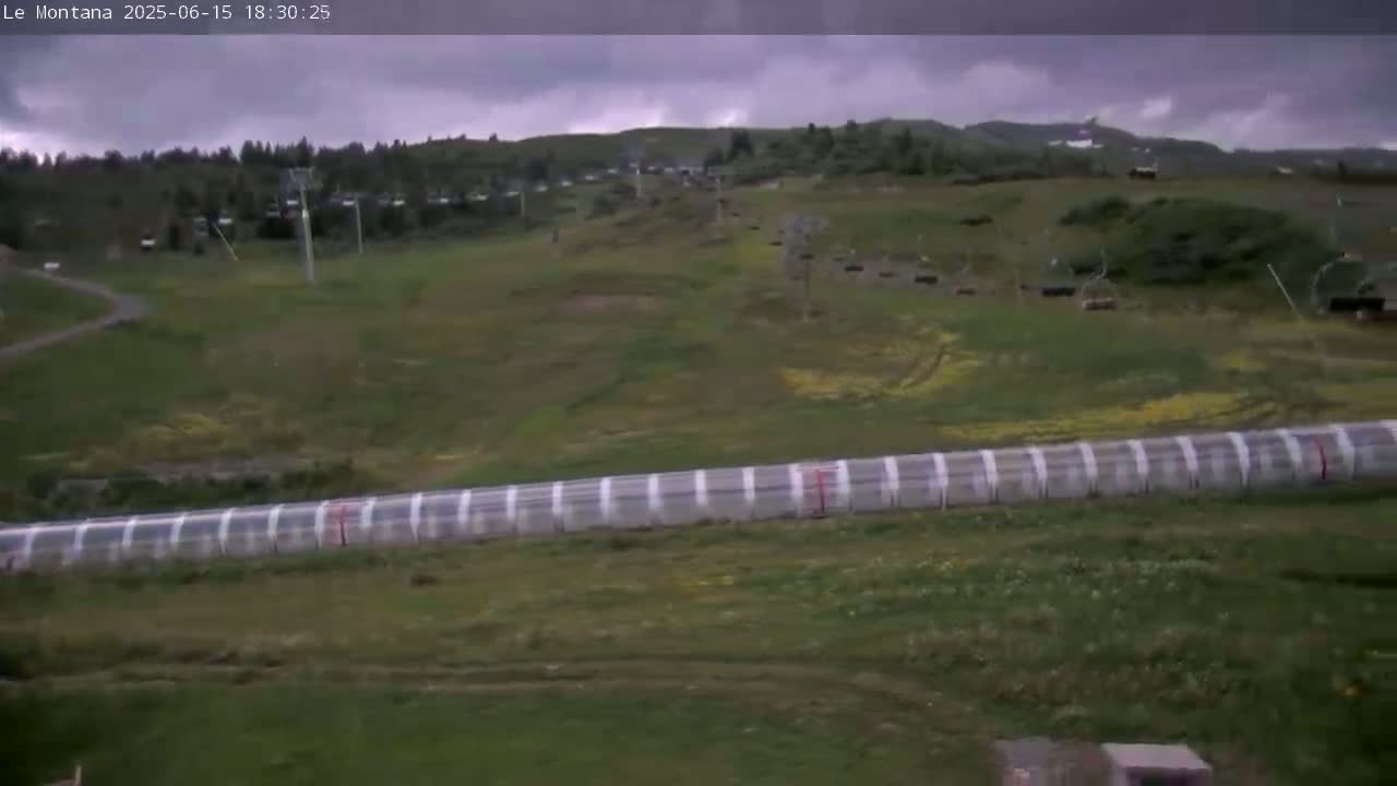 A long, transparent structure is situated on a grassy hillside under an overcast sky, with chairlifts visible in the background.