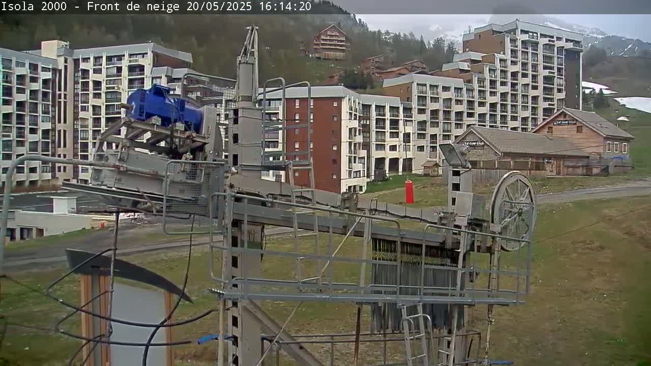 A ski lift mechanism stands in an overcast outdoor setting, with several multi-story buildings and a grassy area visible in the background.