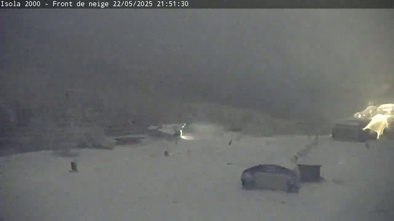 A snow-covered, dimly lit outdoor scene shows a few indistinct structures and vehicles under heavy snowfall.