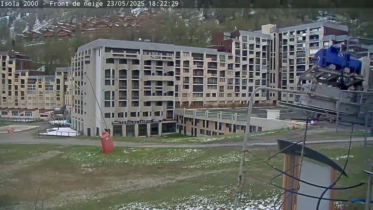 A large apartment building complex sits on a grassy area with patches of snow, viewed from a slightly elevated position, with a ski lift mechanism in the foreground.