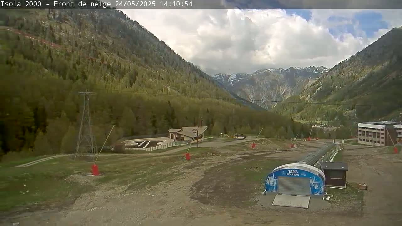 A partly cloudy day reveals a mountainous landscape with green trees, a small building, and a ski lift mechanism in a valley.