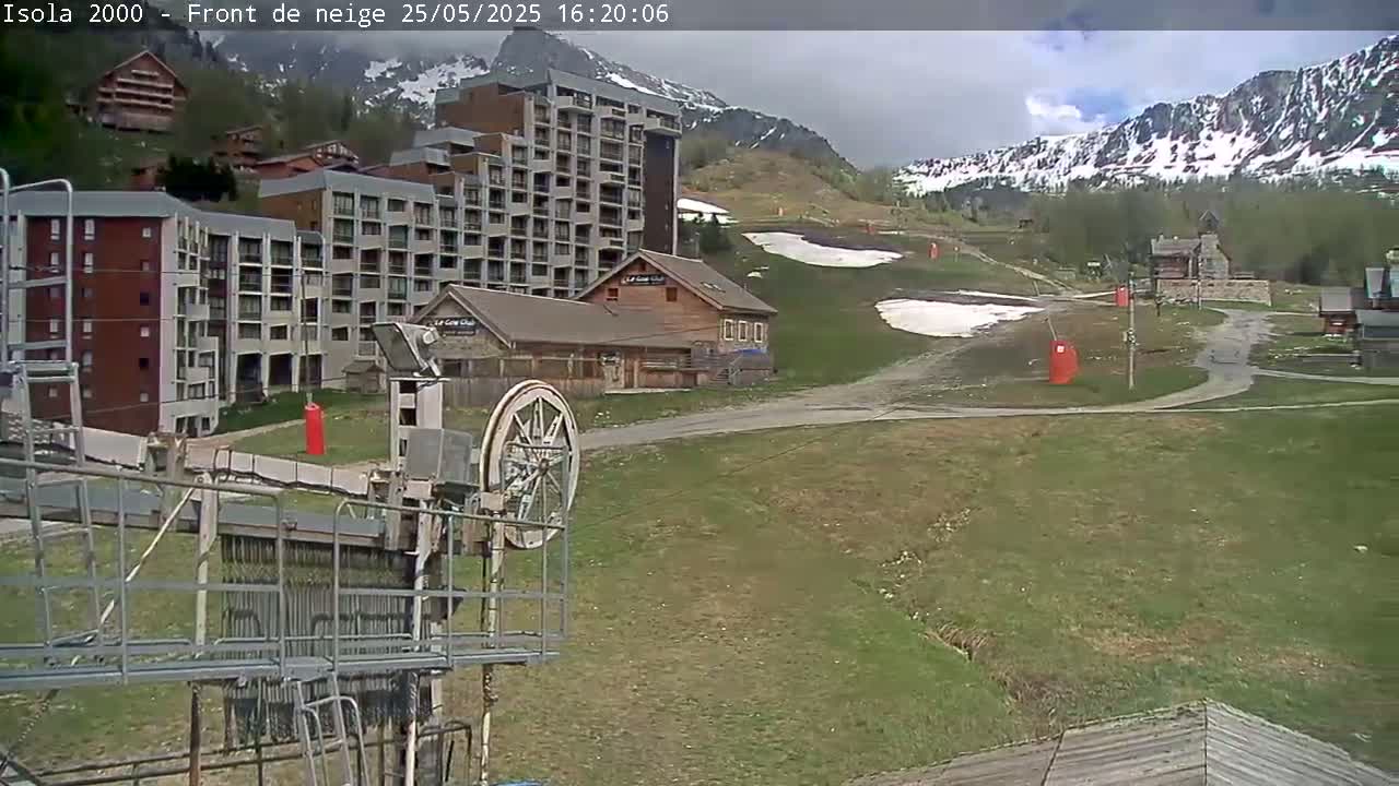 A ski resort with several buildings, a chairlift mechanism in the foreground, and partially snow-covered mountains in the background under partly cloudy skies.