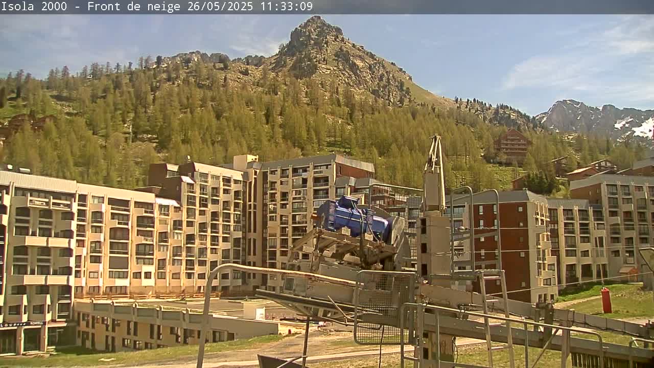 A ski lift and several multi-story buildings are situated at the base of a mountain covered in light green trees under a mostly sunny sky.