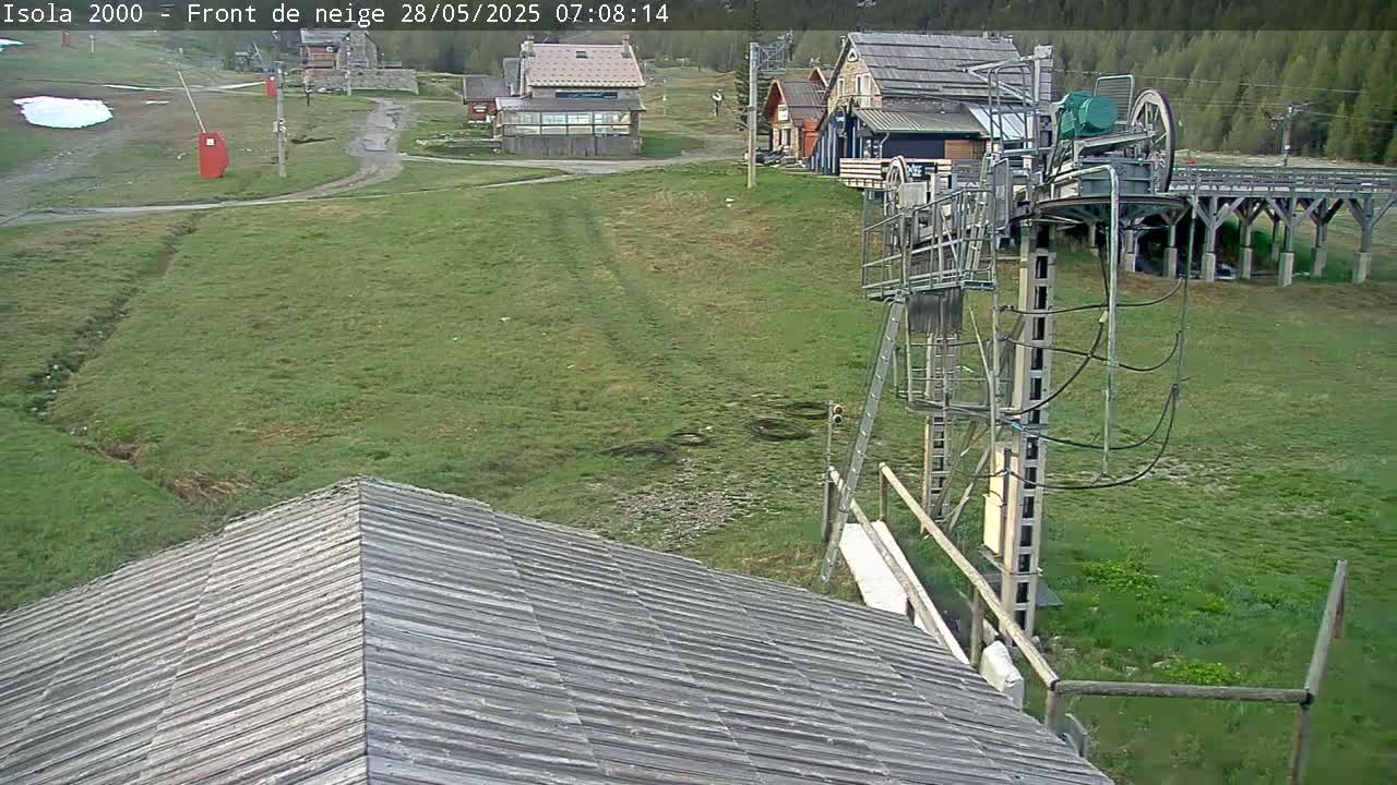 A ski lift stands in a grassy field next to several buildings, under a clear sky.