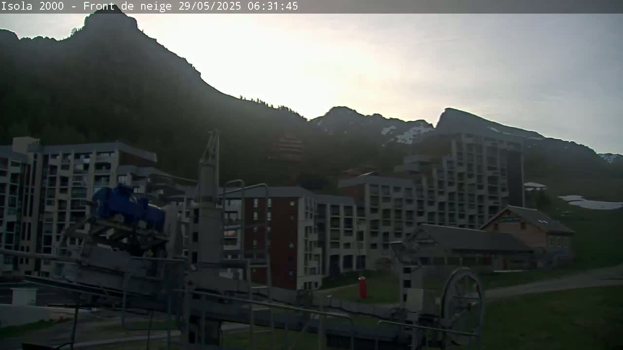 A ski resort at dawn, featuring several multi-story buildings nestled against a mountainside with patches of snow visible in the distance.