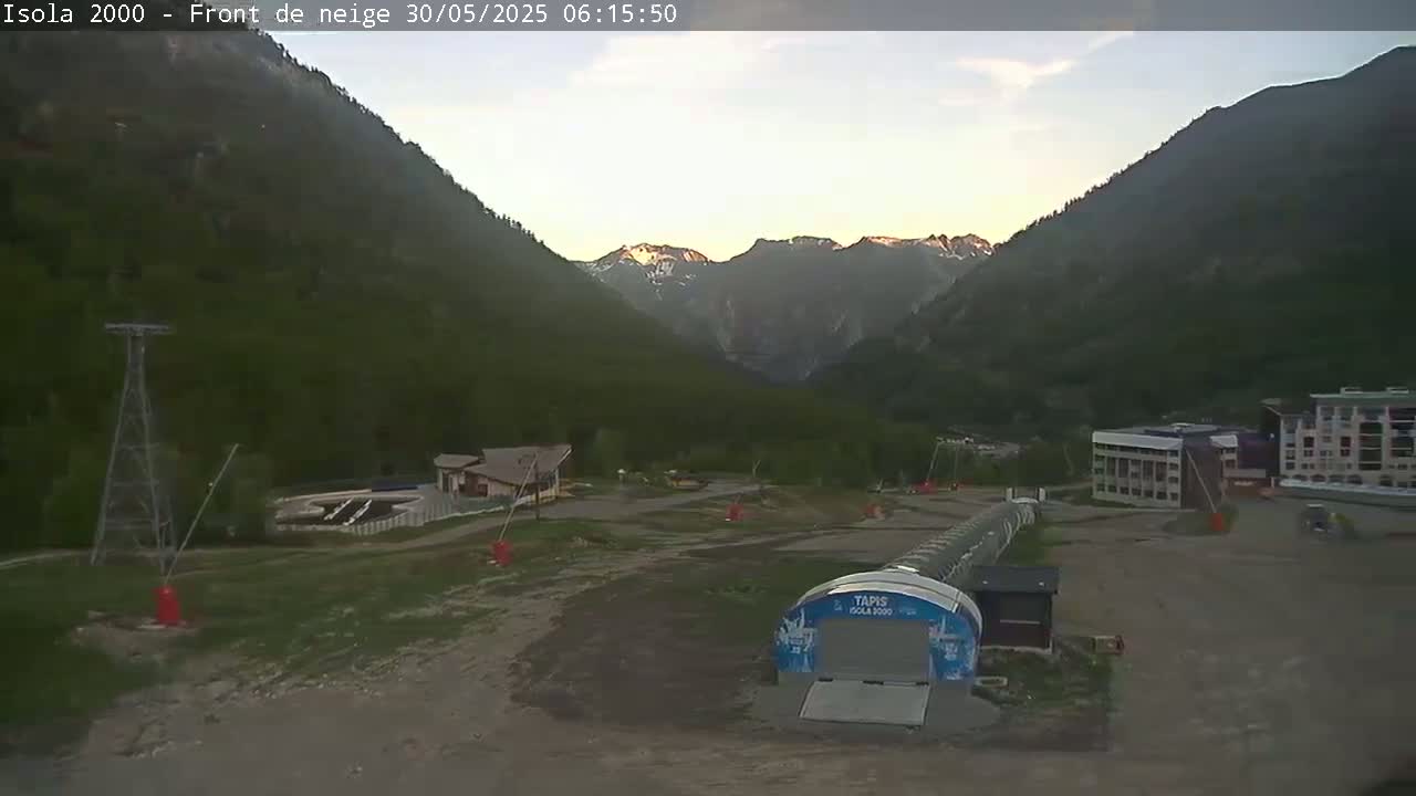 A mountain valley at sunrise shows a ski resort with buildings and a ski lift, under partly cloudy skies.