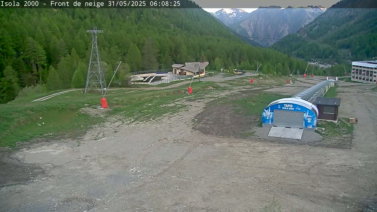 A mostly clear day reveals a mountain valley with a ski lift, a small building, and a covered conveyor lift in a dirt area surrounded by green grass and trees.