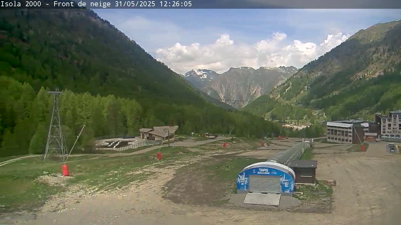 A partly cloudy day in a mountainous region shows a valley with green trees, a few buildings, and a ski lift.