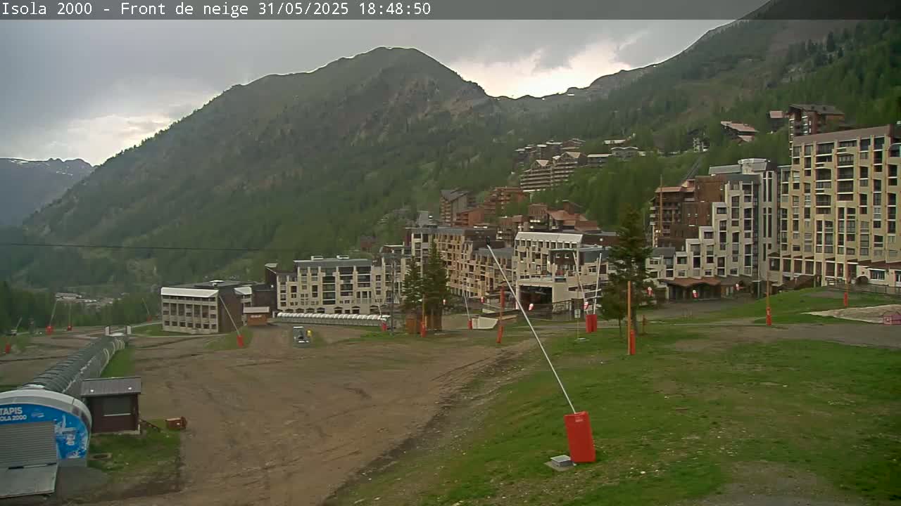 A mountainside village of multi-story buildings under a cloudy sky sits before a large mountain range.
