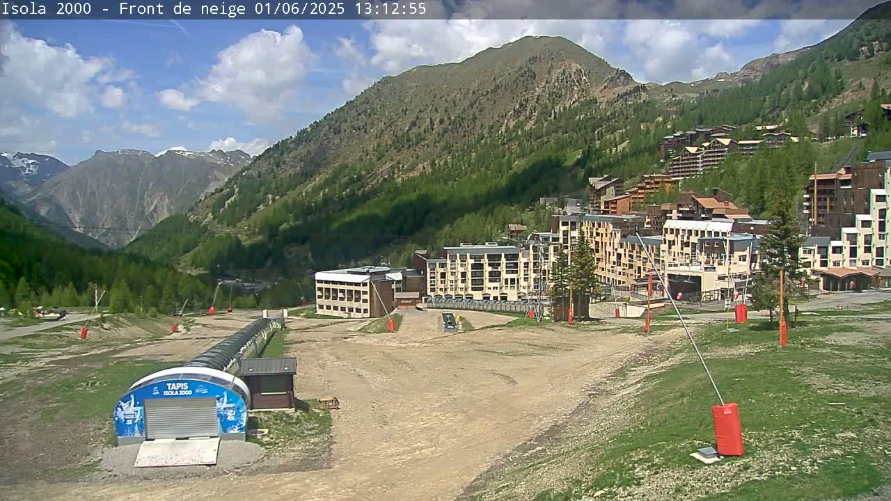 A mostly sunny day reveals a mountainside village nestled in a valley, with several buildings and a ski lift visible in the foreground.