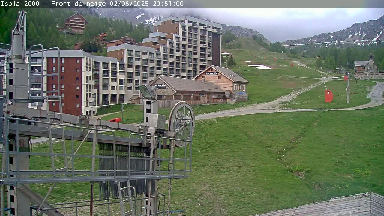 A ski lift pulley system is in the foreground, overlooking a grassy mountainside with several buildings, including apartment complexes and a small chalet, under an overcast sky.