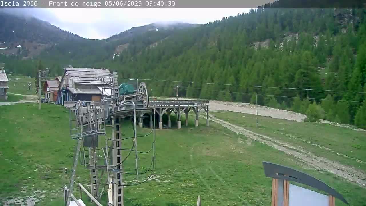 A ski lift and several buildings are situated on a grassy area at the base of a mountain range under an overcast sky.
