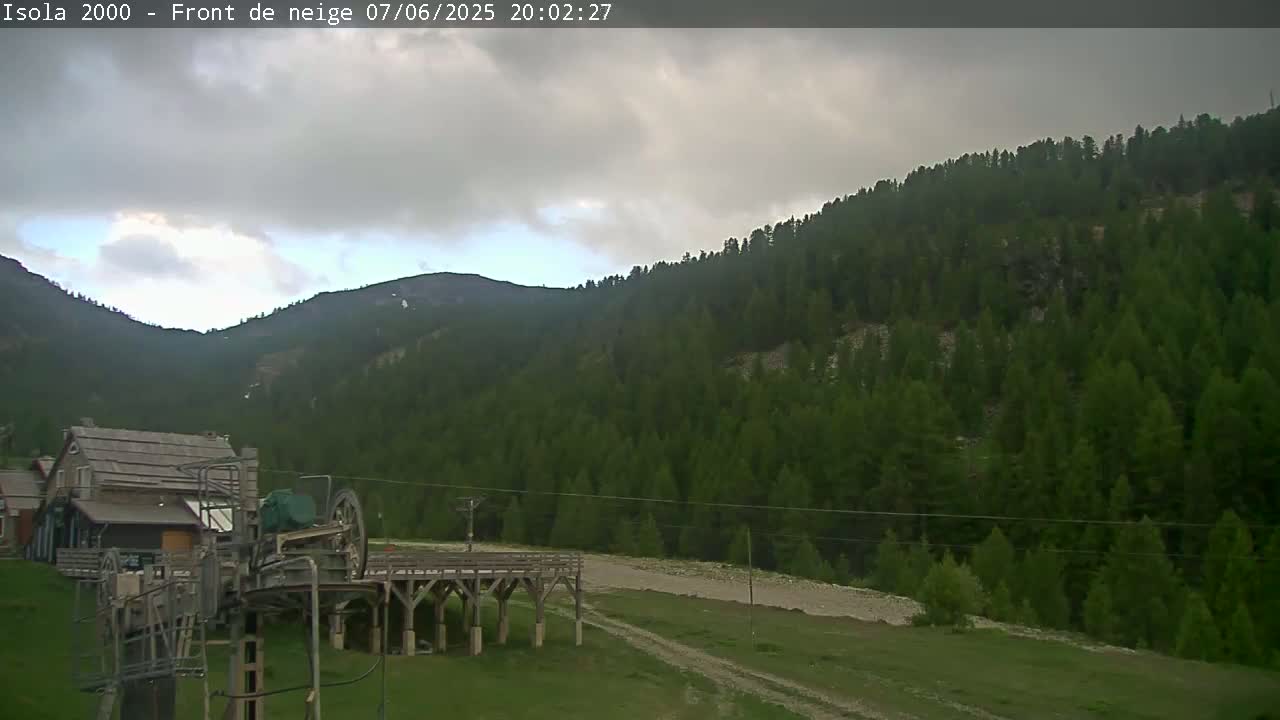 A mostly cloudy sky hangs over a mountain valley covered in dense evergreen trees, where a ski lift and small building are visible in the foreground.