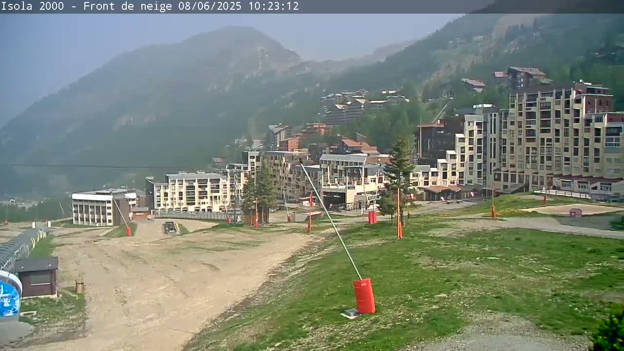 A hazy view of a mountain resort town with several multi-story buildings, a grassy area, and a dirt road in the foreground.