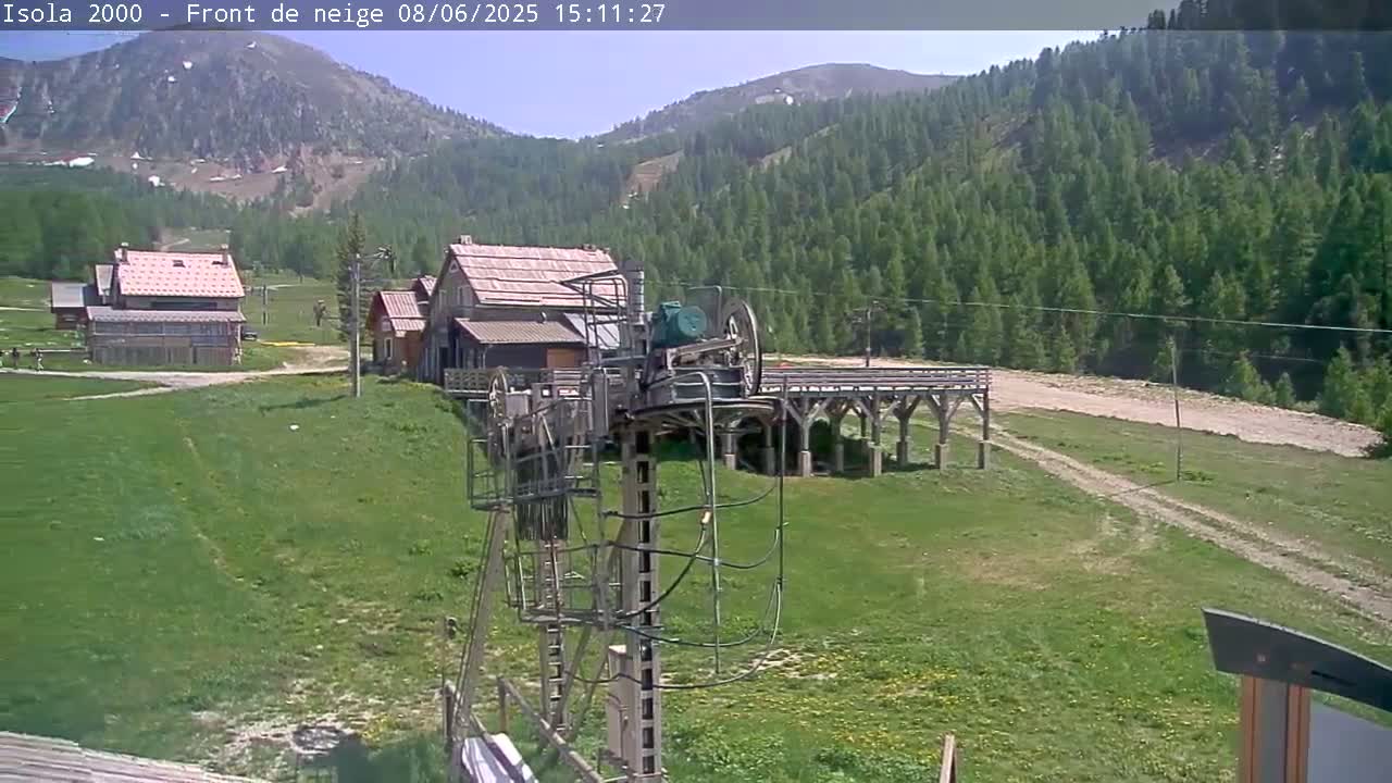 A ski lift mechanism stands in a grassy field, with several buildings and a mountain covered in green trees in the background under a sunny sky.