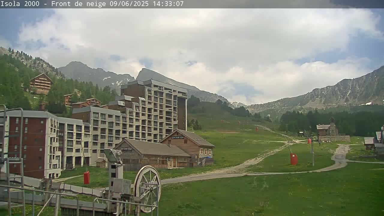 A partly cloudy day reveals a mountain village with several apartment buildings and smaller houses, nestled against a backdrop of large, green-covered mountains.