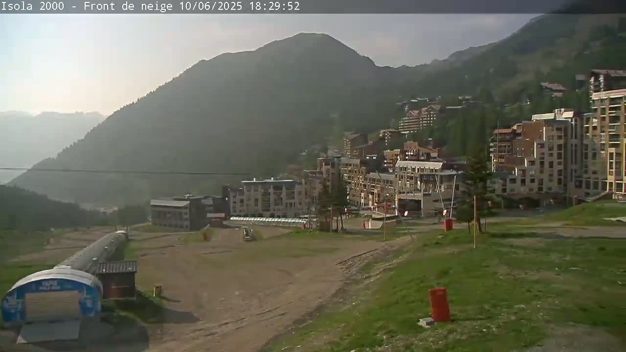 A hazy view of a mountainside village with several multi-story buildings, a grassy area in the foreground, and a long, light-colored structure resembling a pipeline.