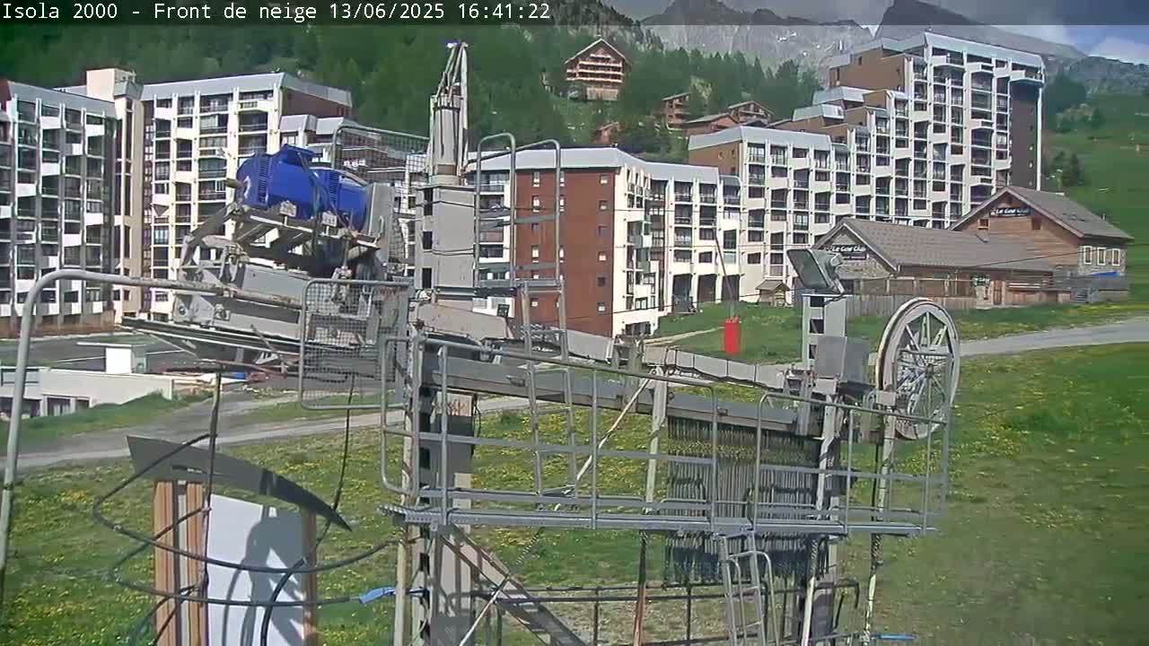 A ski lift mechanism is in the foreground, with apartment buildings and mountains visible in the background on a sunny day.