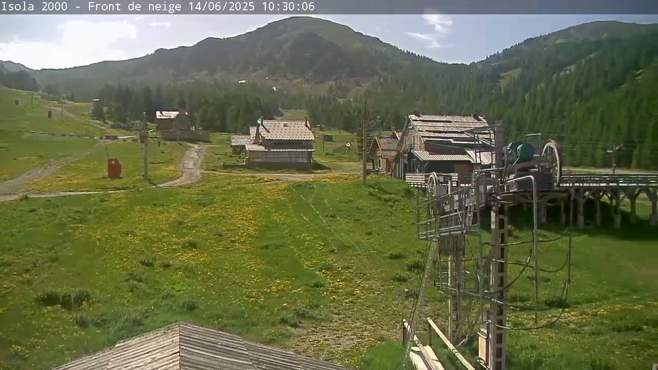 A sunny day reveals a mountainside village with several buildings, a grassy field dotted with yellow flowers, and an old ski lift in the foreground.
