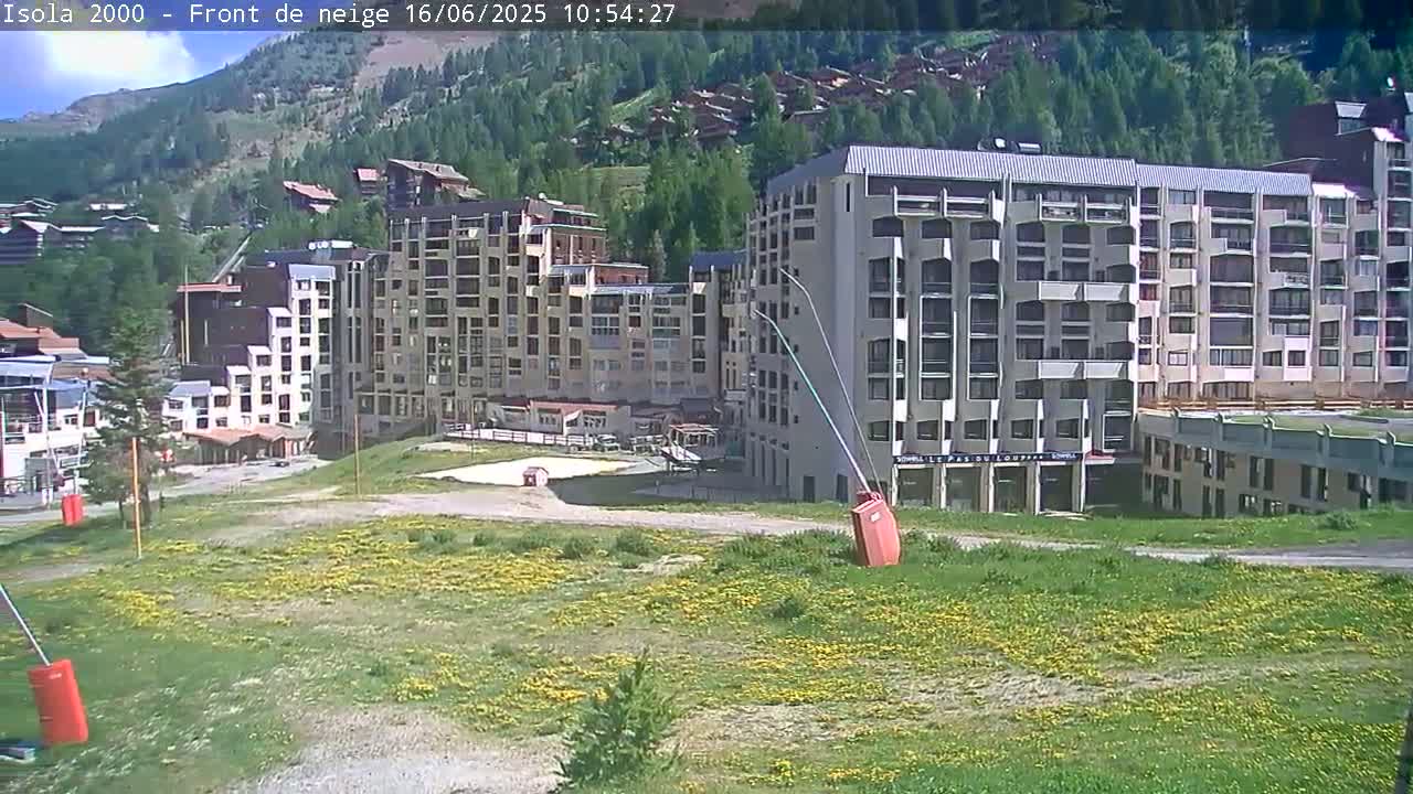 A sunny day reveals a cluster of multi-story buildings nestled in a mountainous region, surrounded by grassy fields dotted with yellow wildflowers.