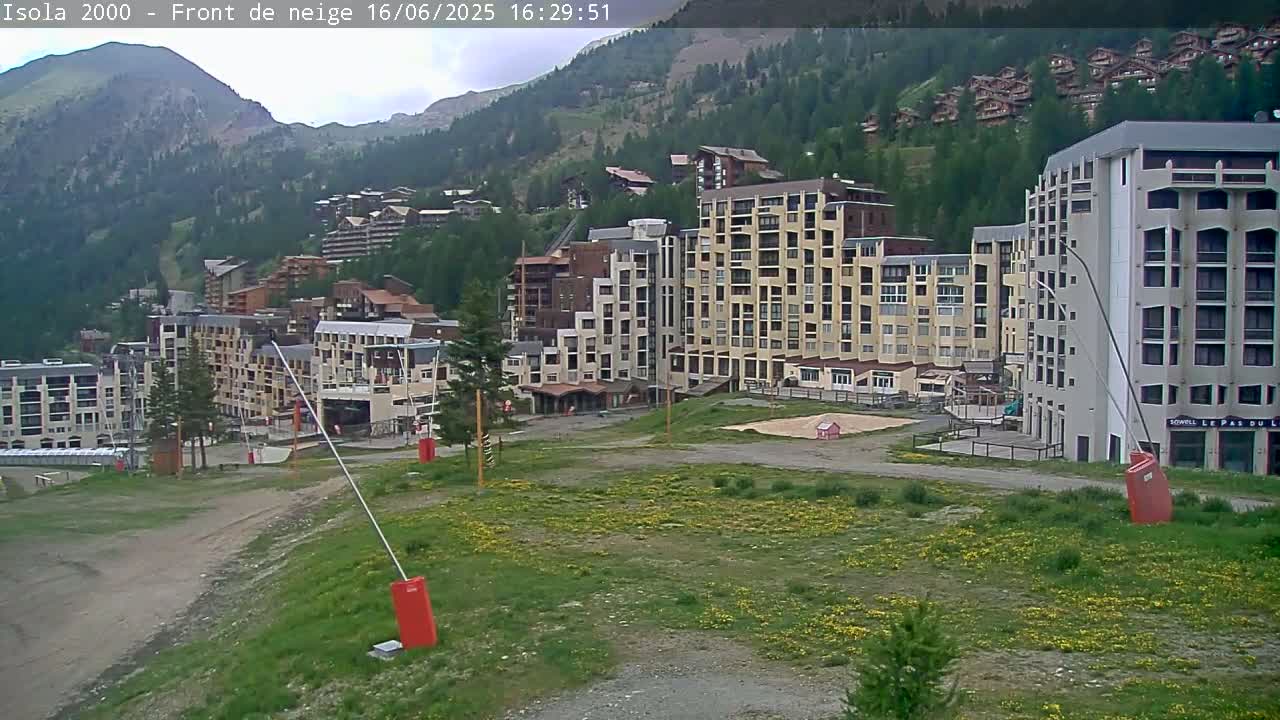 A mountainside village of multi-story buildings under a partly cloudy sky, with grassy areas and ski lift equipment in the foreground.