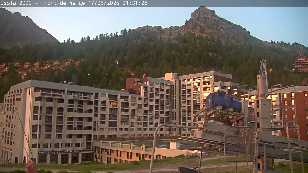 A mountainside village at dusk, featuring several multi-story buildings and a ski lift, with a large mountain in the background.
