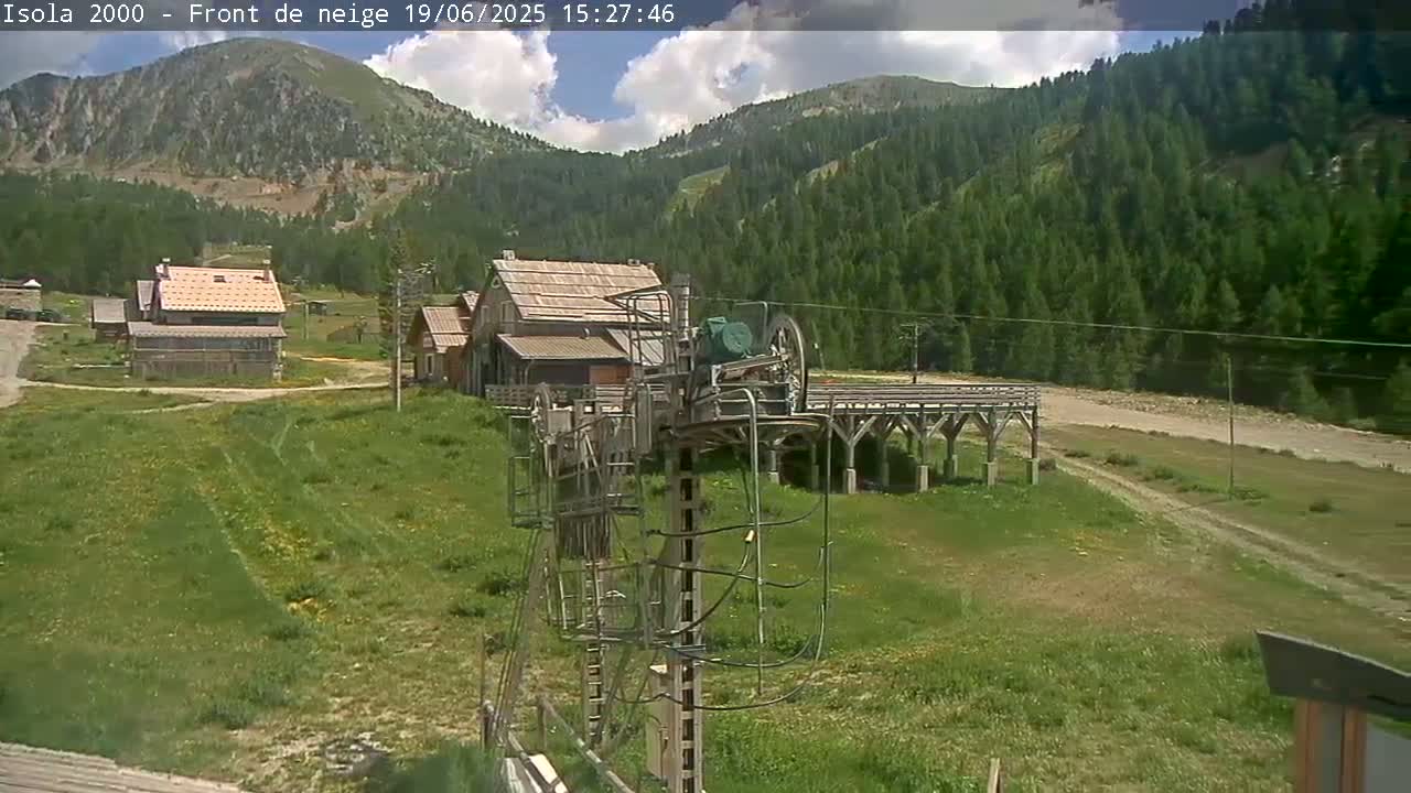 A partly cloudy day shows a mountain ski lift base station with buildings and grassy fields in front of a forested mountain range.