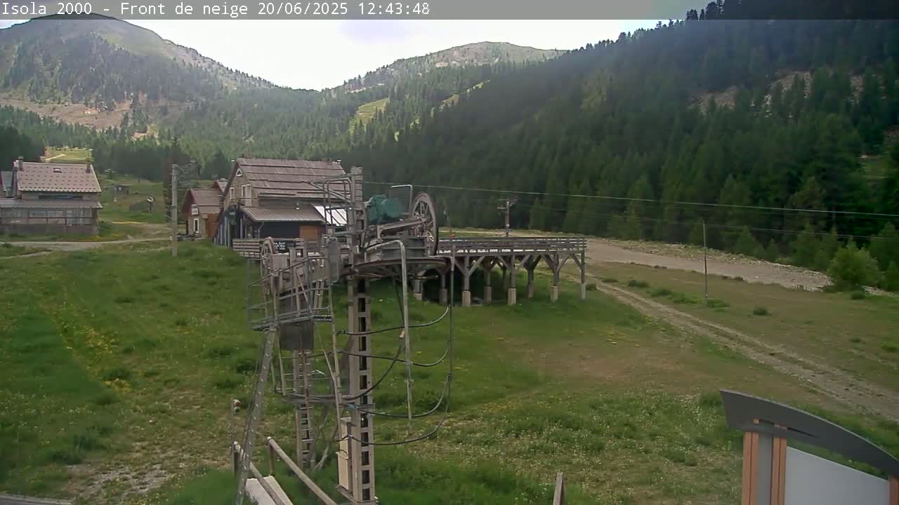 A ski lift sits in a grassy field near several buildings, with a mountain covered in a dense forest visible in the background under a mostly clear sky.