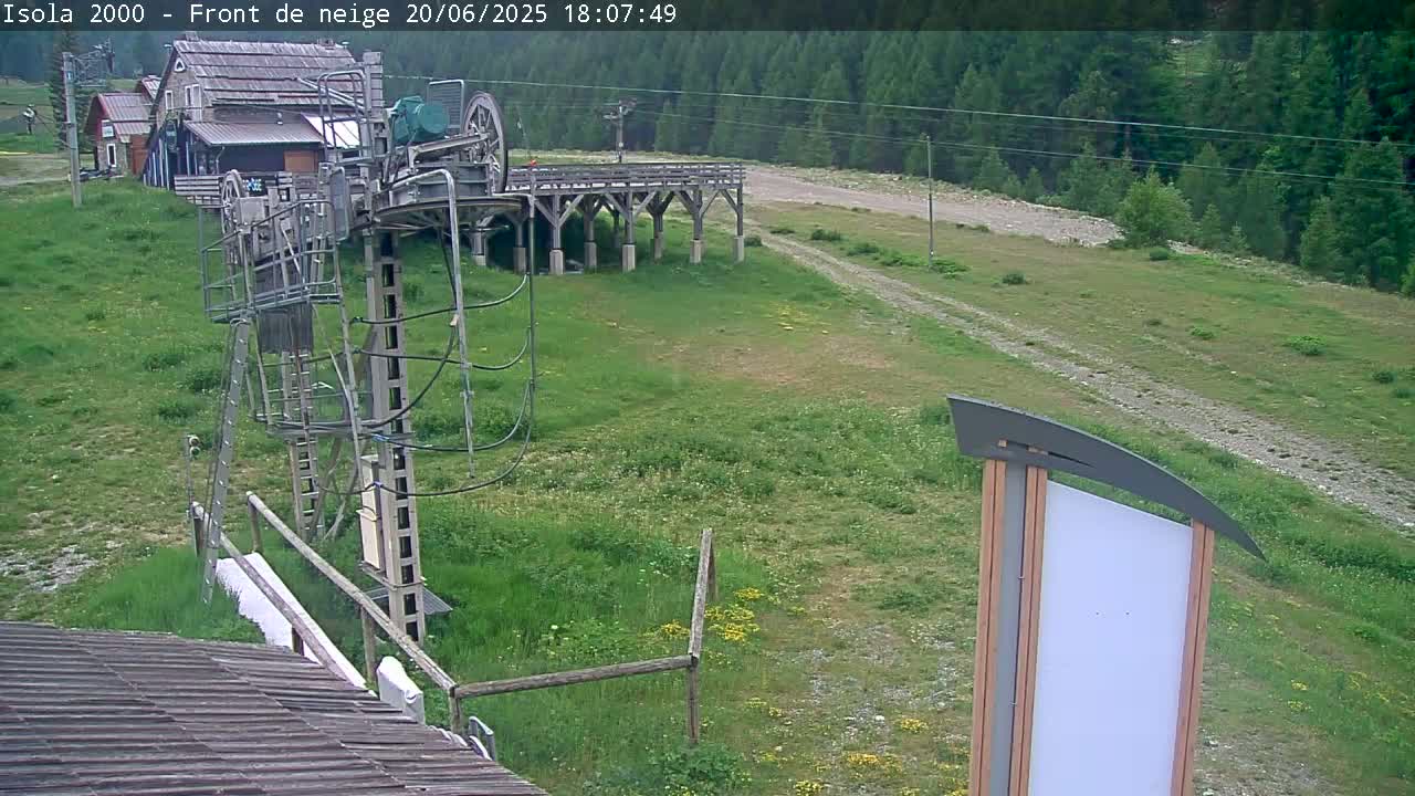 A ski lift mechanism is situated on grassy terrain next to a wooden platform, with a forest in the background under a clear sky.