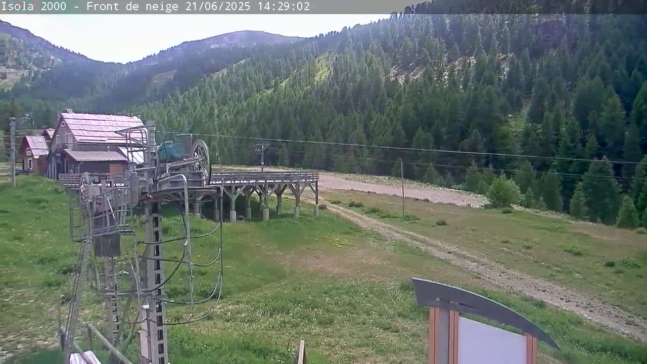 A grassy hillside with a wooden ski lift and small buildings in the foreground, backed by a dense evergreen forest under a mostly clear sky.
