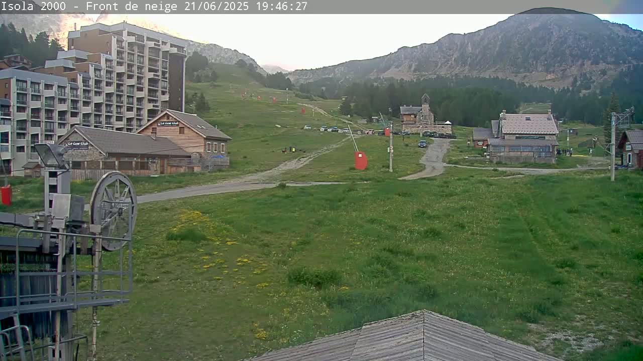 A ski resort in a mountainous area with a mostly clear sky shows a large hotel, several smaller buildings, a grassy area, and a ski lift mechanism in the foreground.