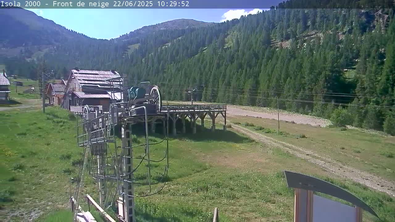 A ski lift base station and several buildings are situated on a grassy area, with a mountain range covered in a dense evergreen forest in the background under partly sunny skies.