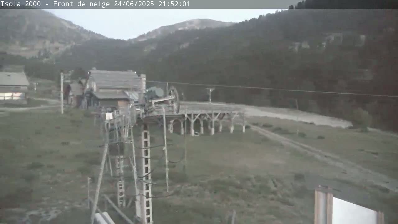 A chairlift mechanism is situated in a mountainous area under overcast skies, near several buildings.
