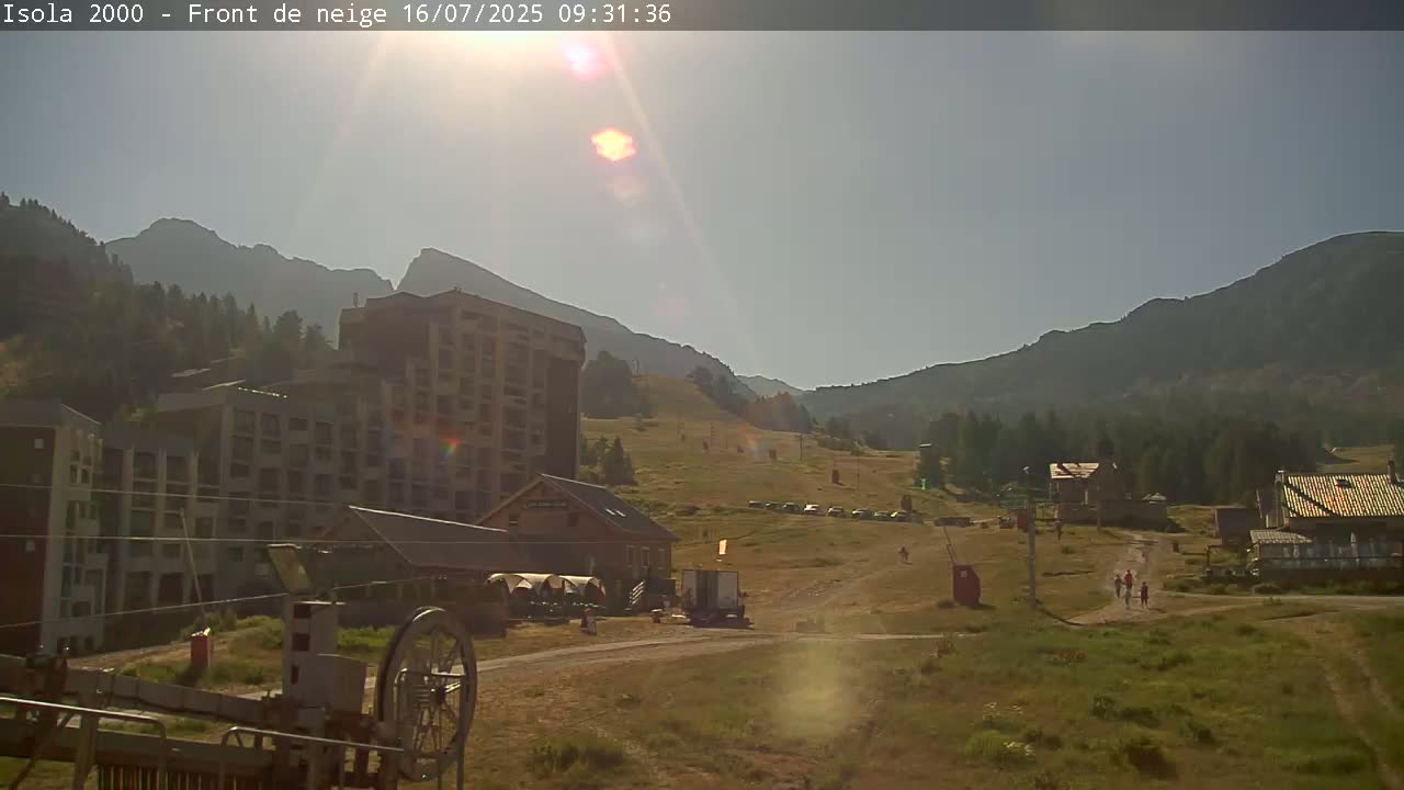 A sunny day in a mountainous region shows several multi-story buildings, a grassy hillside with a few parked cars, and a chairlift pulley system in the foreground.
