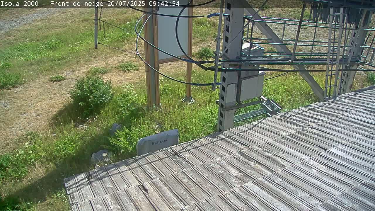 A wooden platform next to a grassy area with a metal structure and wires overhead, on a sunny day.