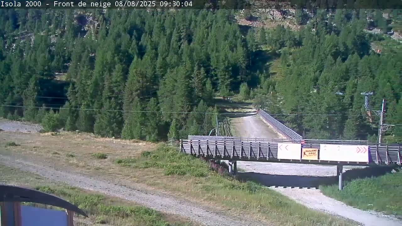 A wooden bridge crosses a dirt road in a sunny, mountainous area filled with evergreen trees.