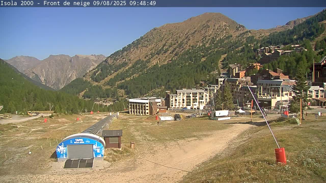 A sunny mountain village scene shows buildings nestled in a valley, surrounded by green hills and mountains under a clear blue sky.