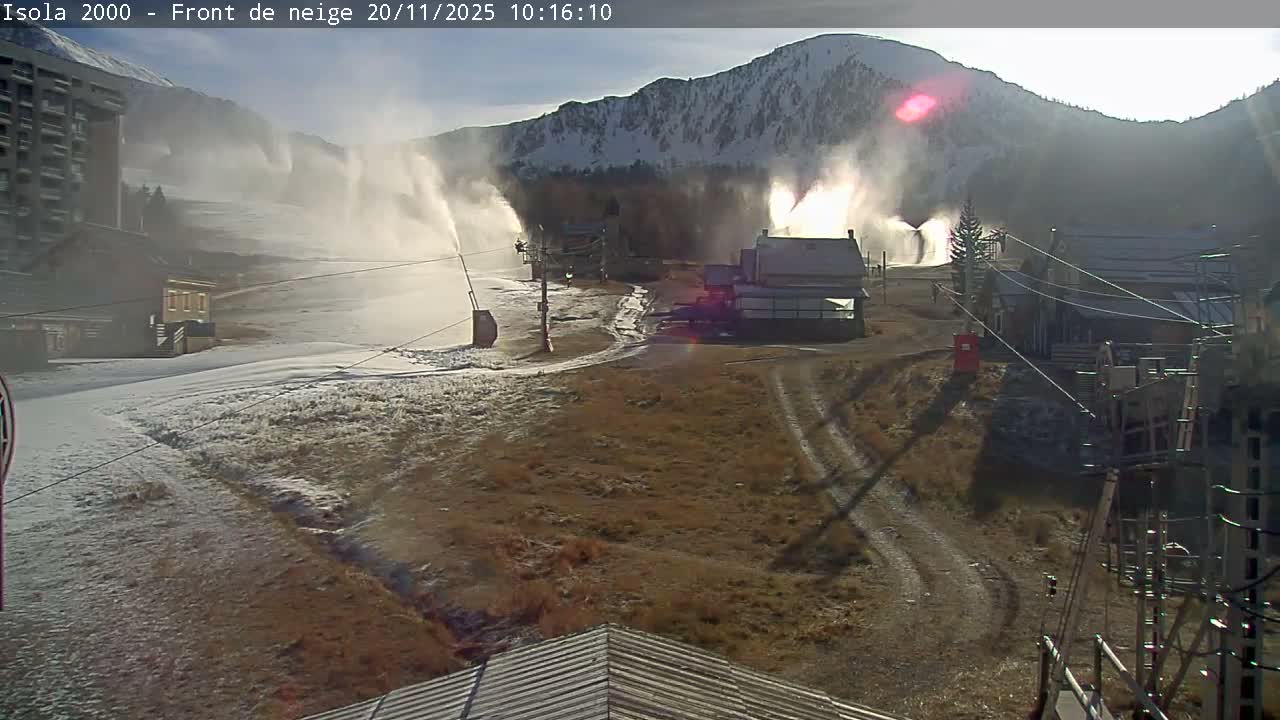 On a sunny and cold day, multiple snow cannons actively blow artificial snow across a partially covered ski slope and adjacent brown terrain, with resort buildings and snow-dusted mountains under a clear sky.