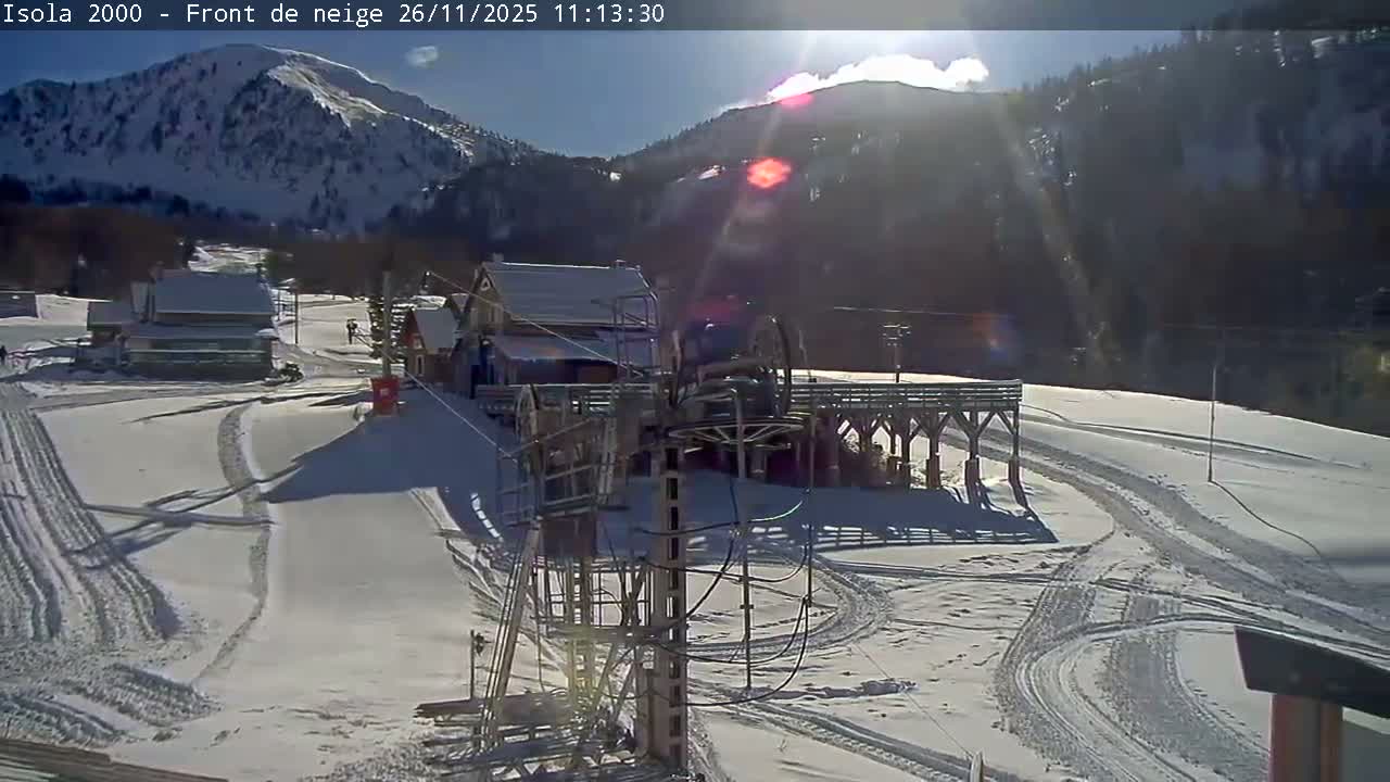 On a clear and sunny winter day, a snow-covered mountain ski resort features multiple buildings with snowy roofs, a prominent ski lift mechanism in the foreground, and numerous ski tracks winding across the slopes, all set against a backdrop of towering snow-capped mountains.
