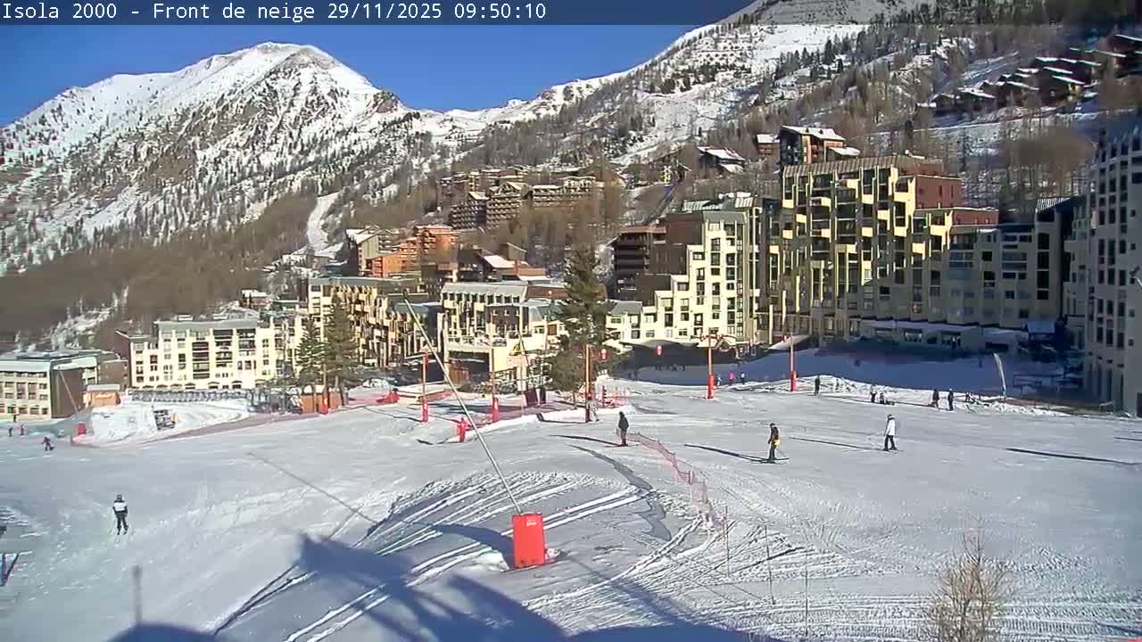 Under a clear blue sky, a lively ski resort base area features skiers on a snow-covered slope, surrounded by numerous multi-story buildings and snow-covered mountains in the background.