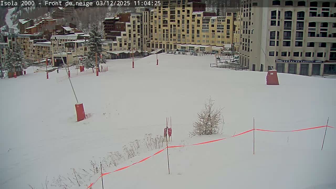 A deserted, snow-covered ski resort area with large buildings and scattered trees is visible, marked by red poles and a barrier tape, all under an overcast winter sky.