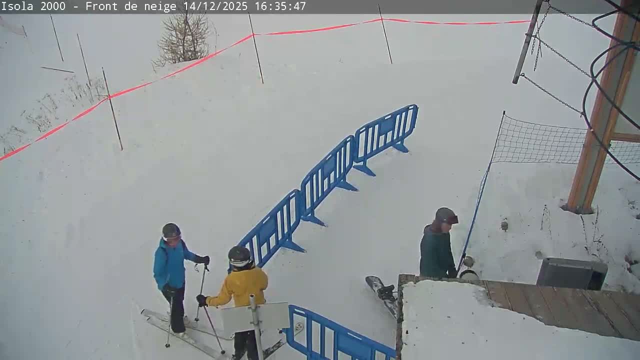 A deserted, snow-covered ski resort area with large buildings and scattered trees is visible, marked by red poles and a barrier tape, all under an overcast winter sky.